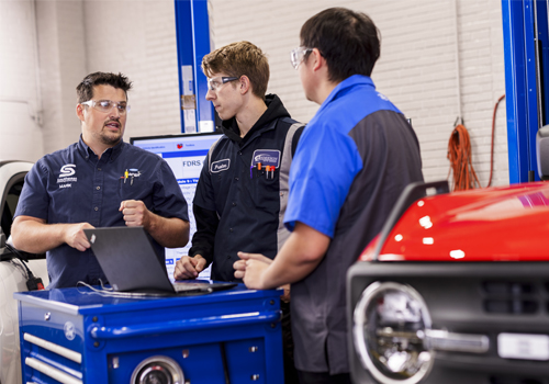 Mark Berggren and two Ford ASSET students are reviewing diagnostic information on a laptop on top of a blue tool cart. Two are wearing dark uniforms with safety glasses, and one is wearing a blue shirt. A red vehicle is partially visible in the foreground, and a hydraulic lift is visible in the background.