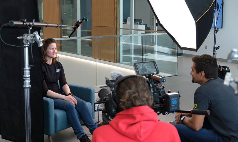 Female in black t-shirt sitting in a chair being interviewed by two men. 