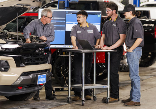 An instructor in the GM ASEP program demonstrates vehicle diagnostics to a group of four technician students in the lab. The instructor stands at the open hood of a tan SUV, gesturing toward the engine while students observe around a mobile diagnostic cart equipped with computer monitors. One additional technician works independently on a black vehicle in the background. All wear dark mechanic uniforms in the well-lit bay with blue tool cabinets visible.