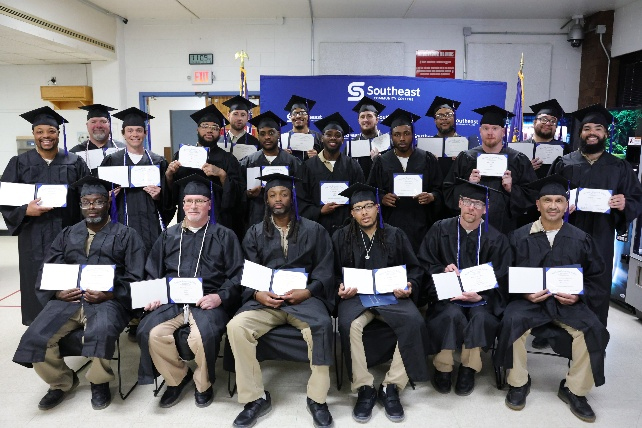 Group photo of more than 20 Southeast Community College graduates celebrating their achievement. Students wearing black graduation caps and gowns are arranged in three rows—one seated and two standing—each holding their certificates. The Southeast Community College blue banner is prominently displayed behind the group in an indoor classroom setting.