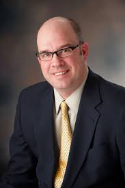 Tim Higgins smiling in a black suit with yellow tie.