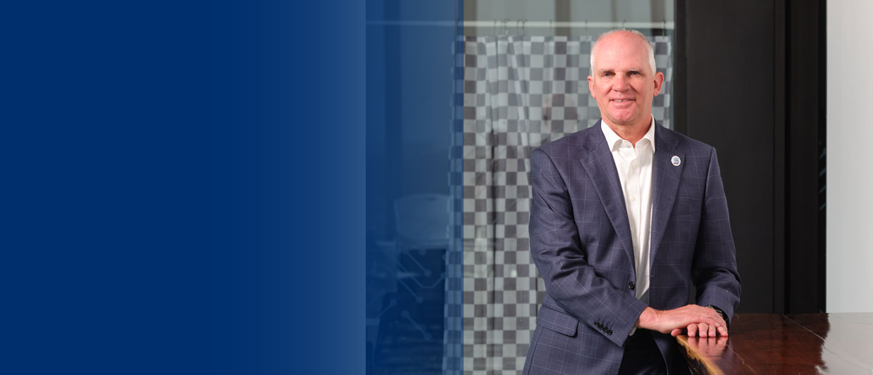 Dr. Paul Illich is in a navy blue suit jacket and white dress shirt, sitting at a table he built for the Sandhills Global Technology Center. The background shows a contemporary office interior with glass and neutral tones.