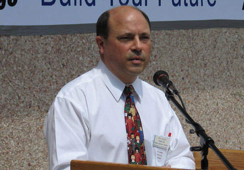 Ron Petsch is in a white dress shirt and colorful patterned tie as he addresses an audience from a wooden podium with a microphone. 