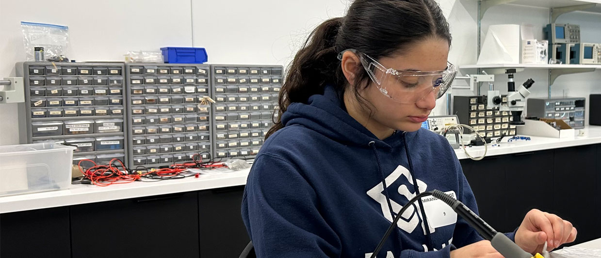 A student wearing safety goggles and a navy blue hoodie carefully uses a soldering iron or precision tool while working at a lab bench.