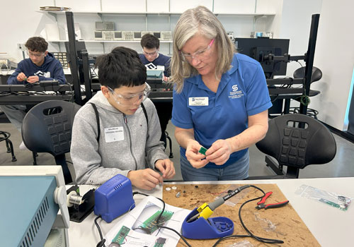 An instructor in a blue shirt assists a young student with hands-on electronics work at a table. 