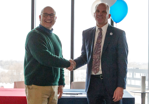 Mike Boehm, smiling in green sweater, and Dr. Paul Illich, smiling in suit, shake hands after signing articulation agreement. 