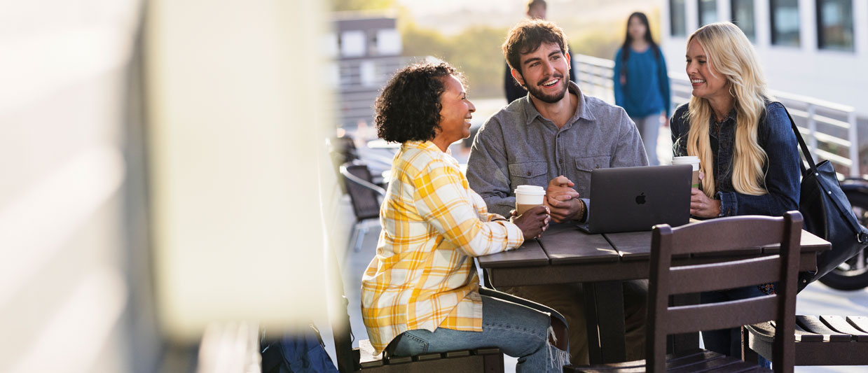 Three students sitting together at an outdoor table on a sunny day, smiling and engaged in conversation. One person has a MacBook laptop open on the table, and all three are holding coffee cups. 