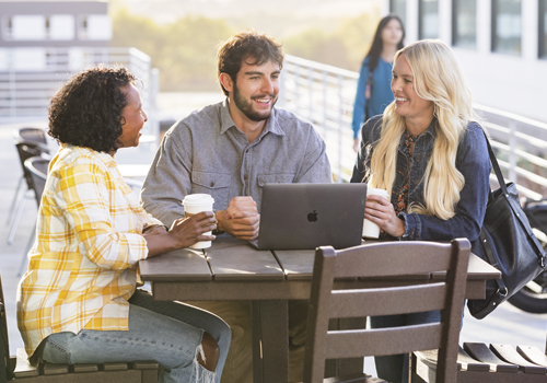 Three students sitting together at an outdoor table on a sunny day, smiling and engaged in conversation. One person has a MacBook laptop open on the table, and all three are holding coffee cups.