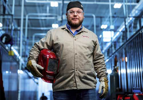 Brayden Kieborz stands in the Welding Technology Center holding a red Lincoln Electric welding helmet and wearing a tan work jacket, safety glasses, and welding gloves. 