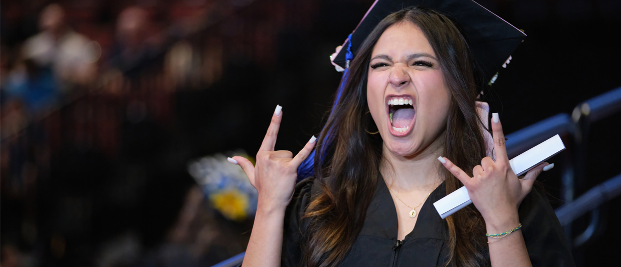 A female graduate in cap and gown celebrates enthusiastically, making rock-on hand gestures and cheering while holding her diploma.