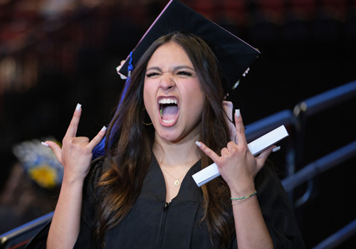 A female graduate in cap and gown celebrates enthusiastically, making rock-on hand gestures and cheering while holding her diploma.