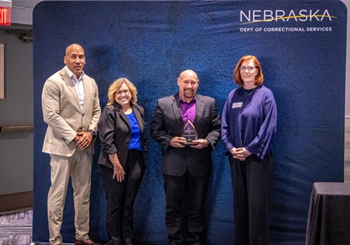 Tommy Moore stands in front of a blue backdrop holding a crystal trophy with a man and woman to the left of him and a woman to the right of him.