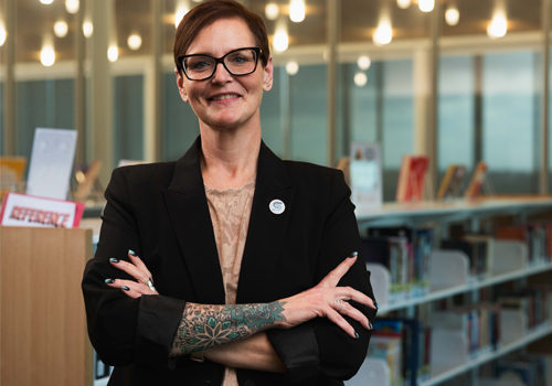 Amy Doty has short brown hair and glasses and is standing with her arms crossed, smiling at the camera. She wears a black blazer over a tan top and has a colorful floral tattoo visible on her forearm. 