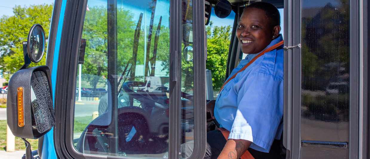 Jessica Whittaker is in a blue Omaha Regional Transit Authority uniform as she sits in the driver’s seat of a blue transit bus, smiling and looking over her shoulder through the open window. 