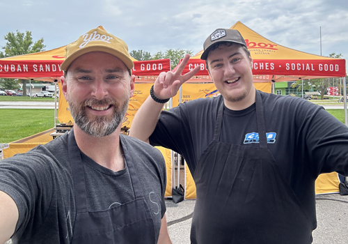 Bill Radtke (left) takes a selfie with a Ybor chef at a pop-up. Radtke is wearing a yellow cap and gray t-shirt while the chef is wearing a dark cap and t-shirt giving a peace sign. Both men are smiling. 
