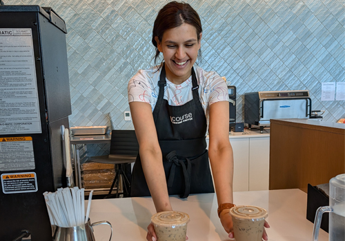 A smiling barista in a black apron sets two iced coffees in lidded plastic cups on a white counter in a bright café with blue-gray tiled walls.