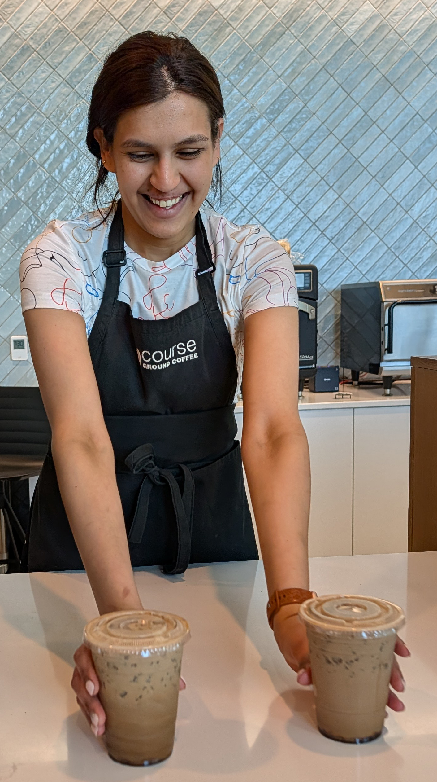 A smiling barista in a black apron sets two iced coffees in lidded plastic cups on a white counter in a bright café with blue-gray tiled walls.