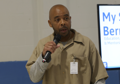 Bernard Long shares his presentation at the UPWARD Scholars Conference. He is wearing a khaki uniform shirt with an ID badge. He has a shaved head and short beard. 