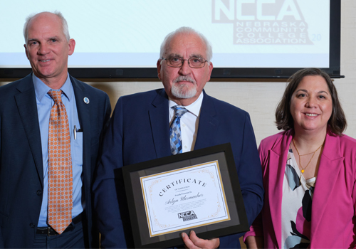 Three business professionals pose for a photo at an awards ceremony. On the left, a person in a blue suit jacket and orange tie; in the center, a person in a navy suit holding a framed certificate; and on the right, a person in a bright pink blazer. 
