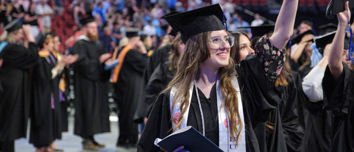A female graduate in cap and gown waves at her family. 