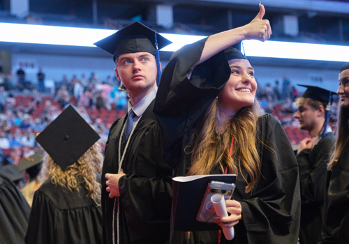 Female graduate with long blonde hair gives a thumbs up as a male graduate looks on. 