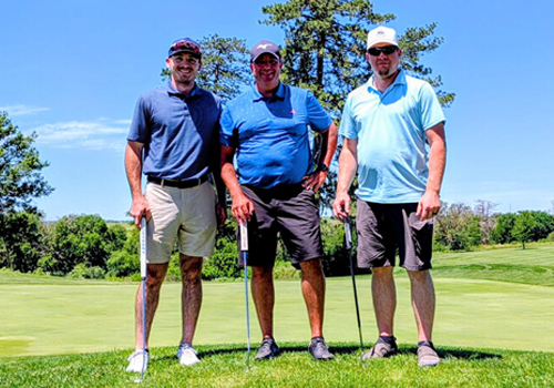 Members of the Cheever Construction team pose at Pioneers Golf Course holding their putters. They’re standing on a green with trees and a fairway in the background on a sunny day.