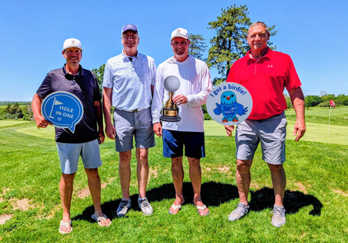 Members of the HVAC Kings team pose at Pioneers Golf Course, holding novelty signs: "Hole in One,” "I Got a Birdie," and a trophy. They're standing on a green with trees and a fairway in the background on a sunny day.